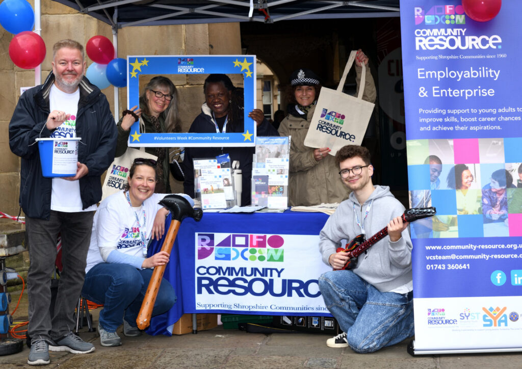Community Resource team posing for a group picture around their stand, all holding various props such as inflatable hammers and tote bags, at the volunteers in the square event.